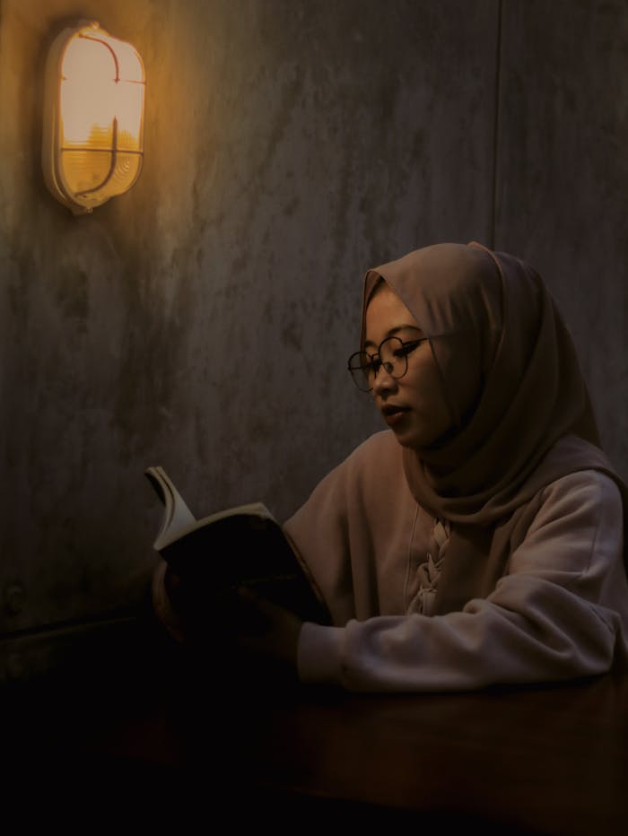 A Muslim woman in a hijab reads a book under warm indoor lighting, creating a serene and thoughtful atmosphere.