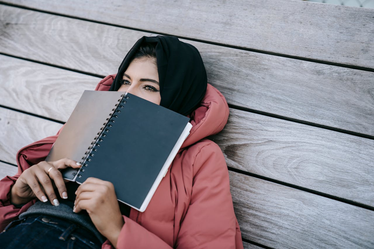 High angle of young Middle East female in casual clothes and traditional headscarf lying on wooden floor of terrace and covering face with notebook