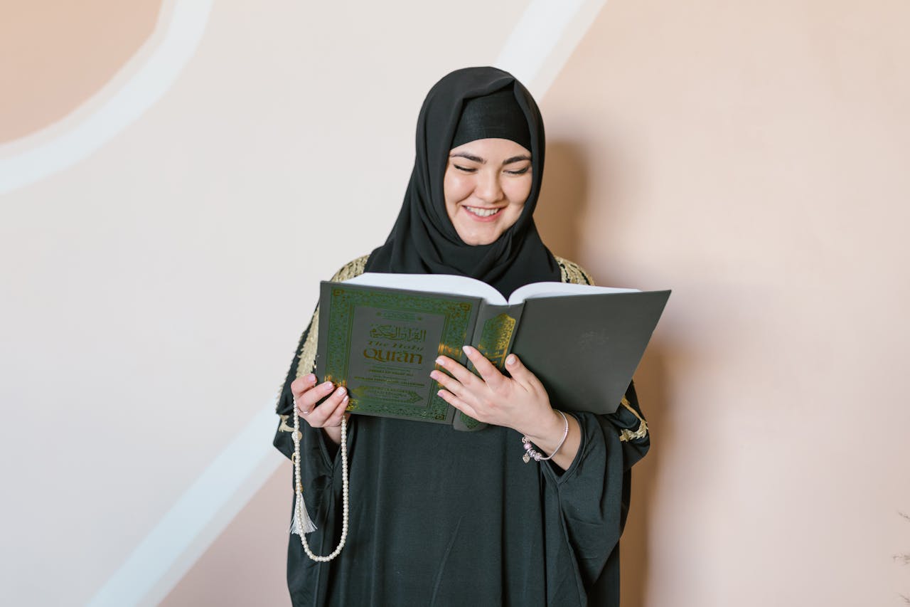 A woman in a hijab, smiling while reading the Quran indoors, embodies faith and tradition.