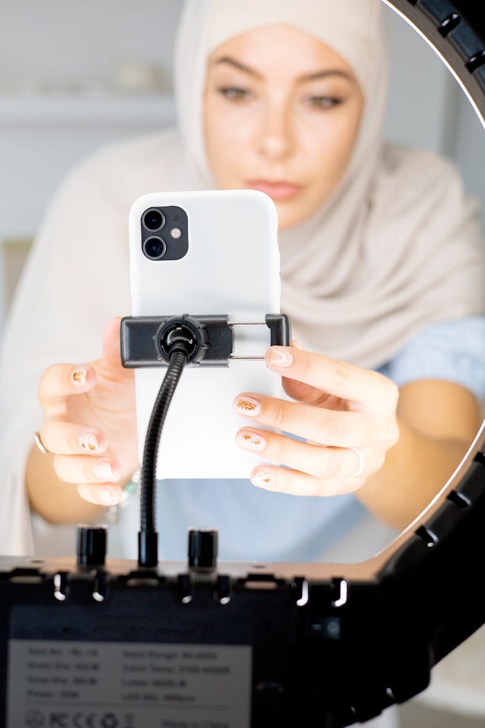 Muslim woman in hijab recording video with smartphone and ring light indoors.